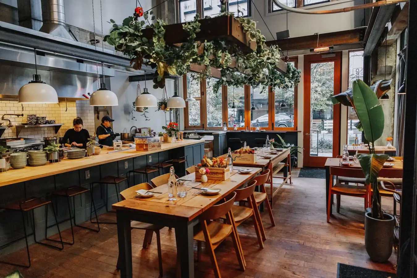 Xóm dining room — warm wooden tables set for dinner beneath hanging greenery, open kitchen in the background