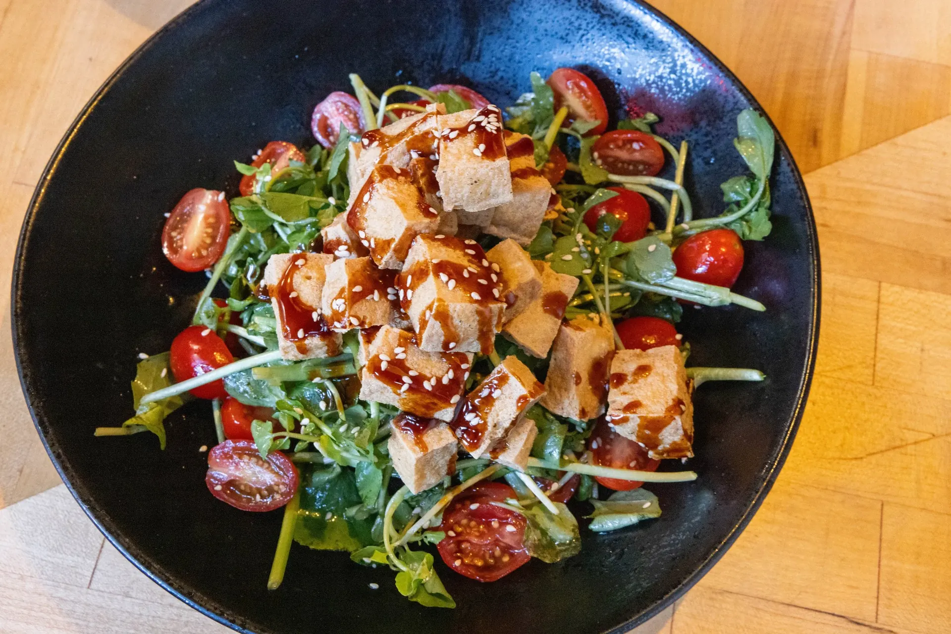 Tofu salad in a dark ceramic bowl with arugula, cherry tomatoes, and sesame dressing