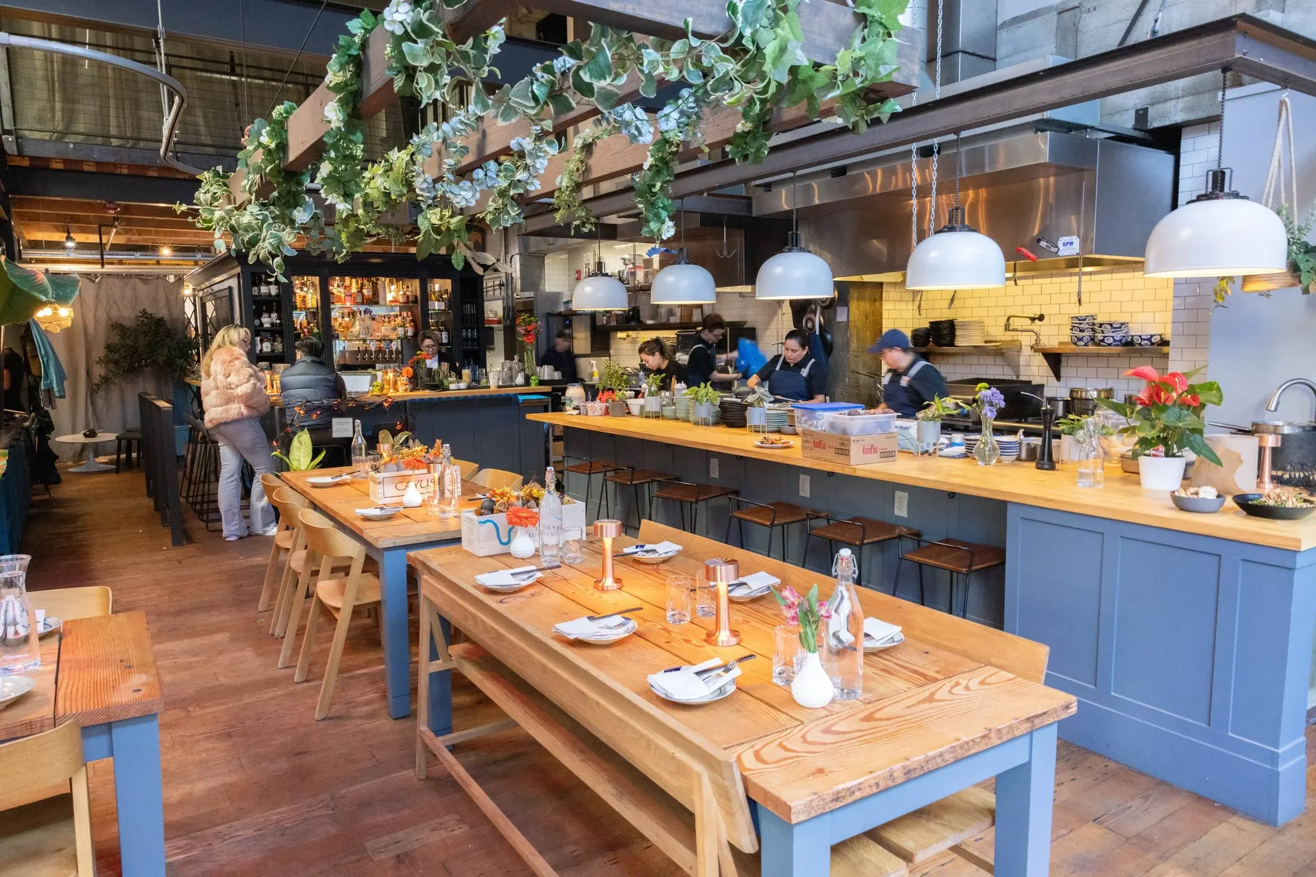 Xóm interior — long wooden tables, hanging greenery, and the open kitchen in the background