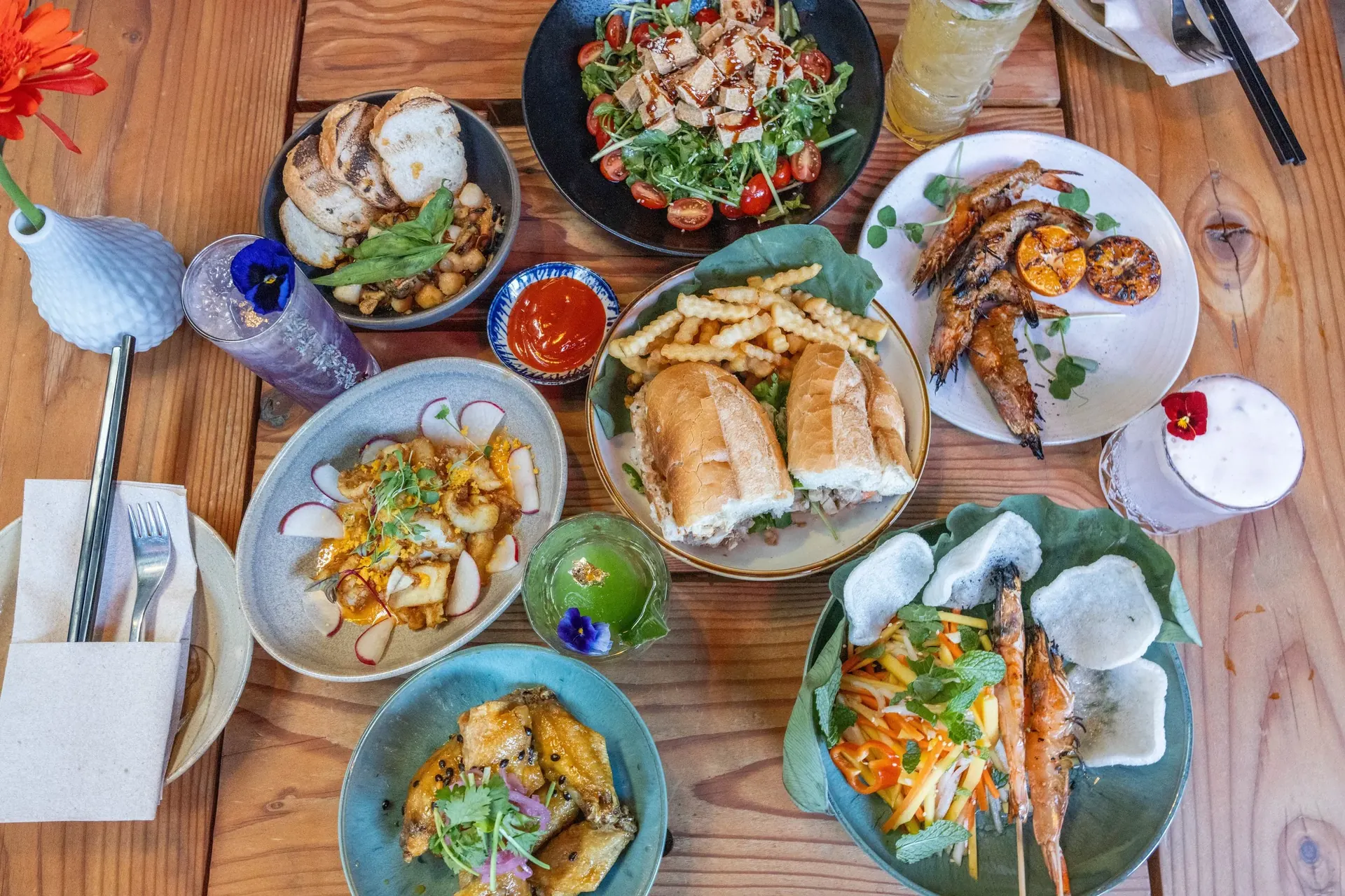 An overhead spread of Xóm dishes — spring rolls, tiger prawns, bánh mì, and cocktails across a wooden table