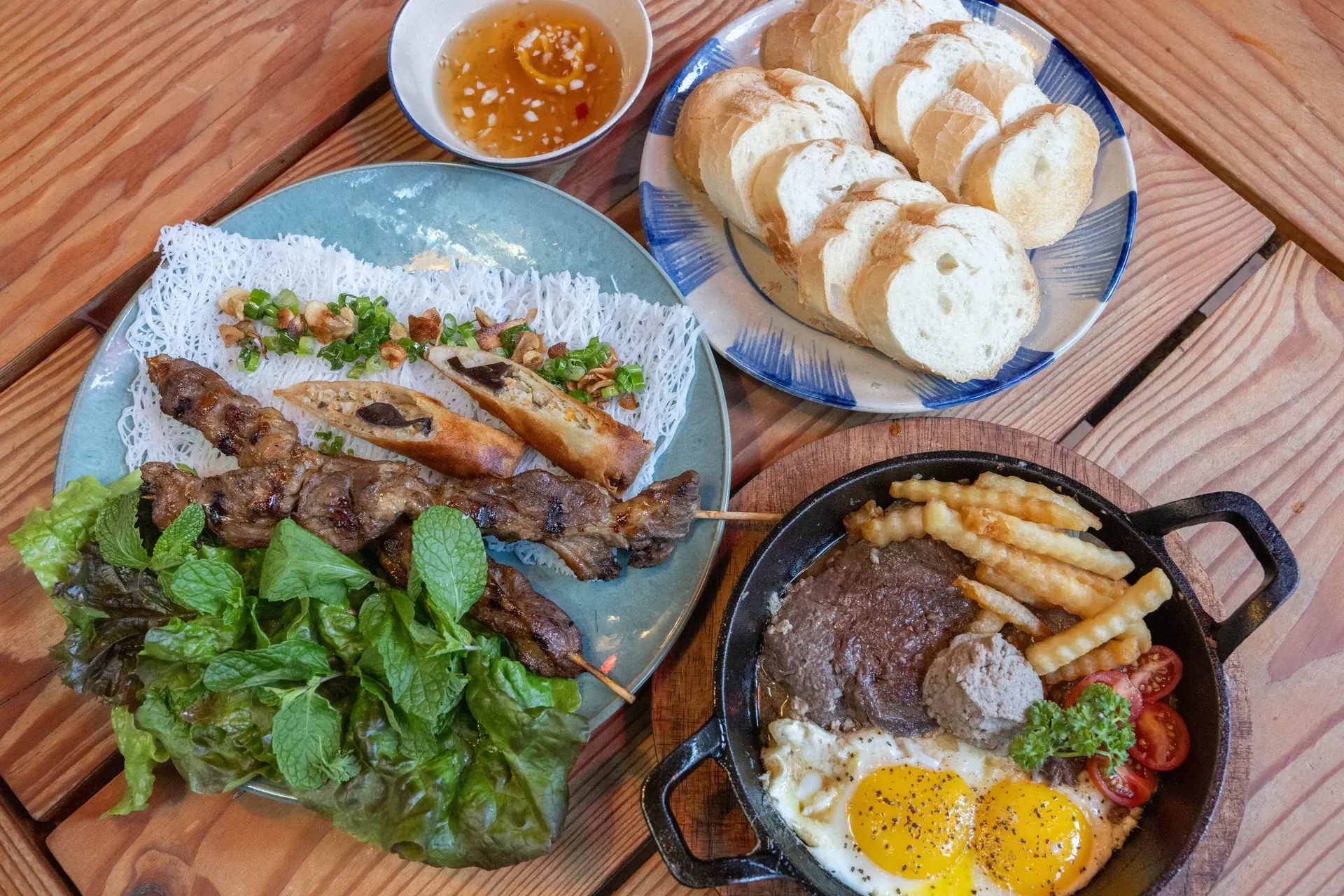Overhead view of Xóm dishes including grilled skewers, Bò Né, and bread